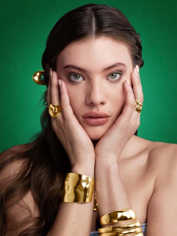 Person on a green background wearing various pieces of gold jewelry including two rings, a bracelet, and bangles.