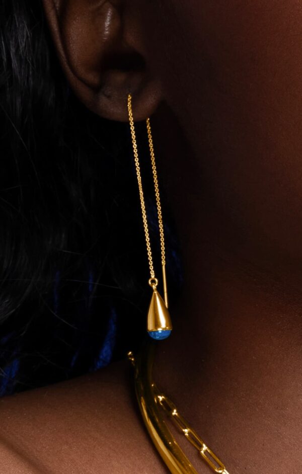 A gold earring with long chain tops and droplet shaped blue bottoms in someone's left ear.