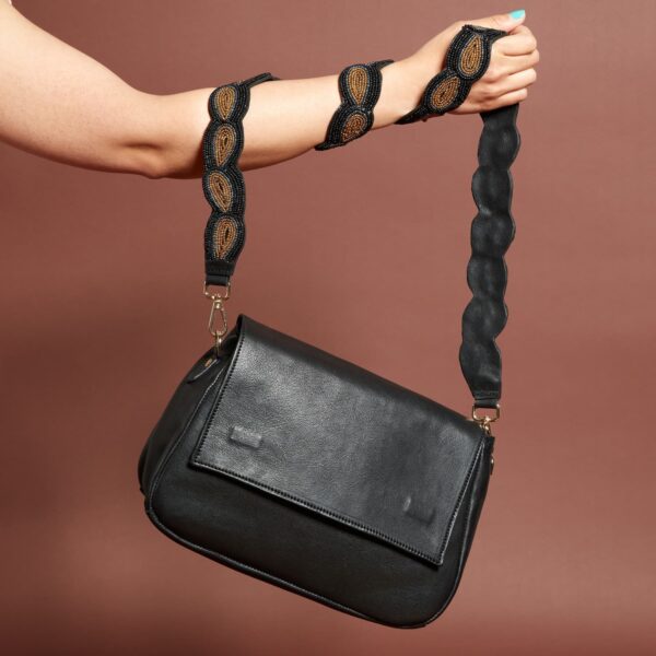 A black leather handbag with a front flap and a beaded brown and black shoulder strap. A light skinned arm is holding the bag with the strap wrapped around the forearm a few times.